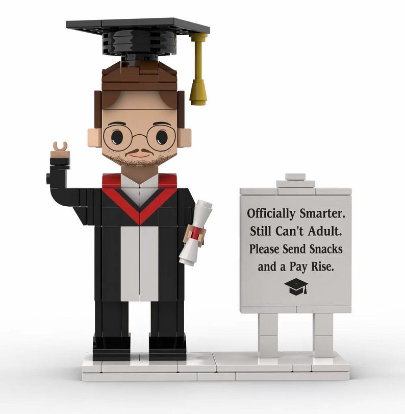 May include: A graduation-themed figurine made of toy blocks. The figure wears a black graduation gown with red trim, a mortarboard, and glasses. It holds a diploma and stands next to a sign that reads, "Officially Smarter. Still Can't Adult. Please Send Snacks and a Pay Rise."