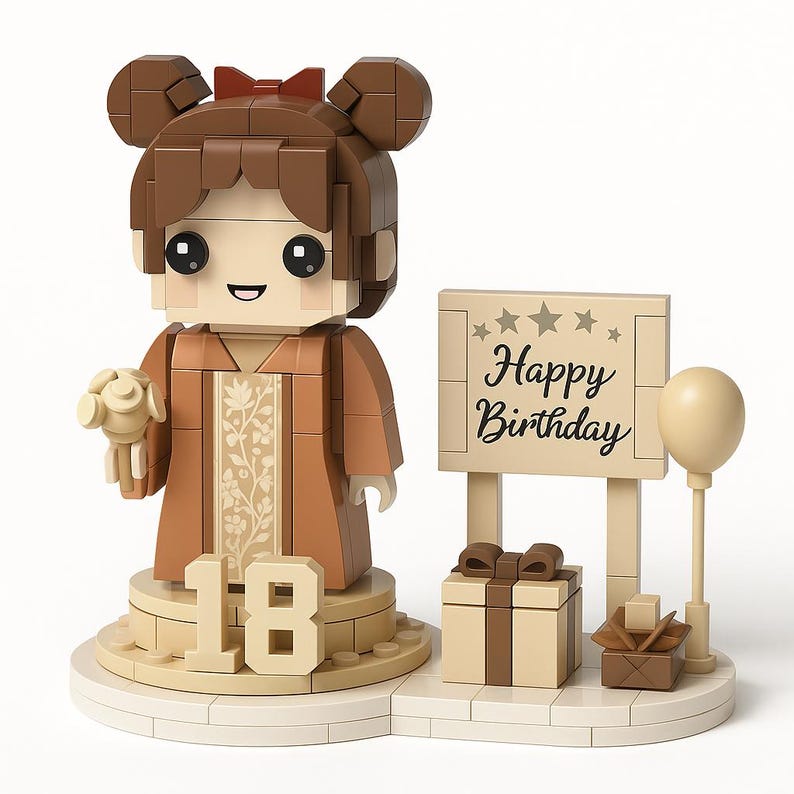 May include: A brown and beige Lego-style birthday figurine. The figurine features a girl holding a bouquet, with the number 18 at the base. A sign that says "Happy Birthday" is next to a gift box and a balloon.