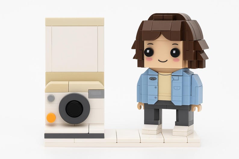 May include: A Lego brick-built figure of a person wearing a blue jacket and dark pants, standing next to a Lego brick-built camera. The camera has a black lens and an orange button. The figure has brown hair and a smiling face.