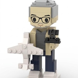 Peut inclure: Figurine Lego représentant une personne avec des lunettes, un chapeau gris et une veste beige, tenant un avion blanc et un objet noir. La figurine est sur une base blanche. La personne a une barbe grise et porte une chemise grise et un pantalon bleu marine.