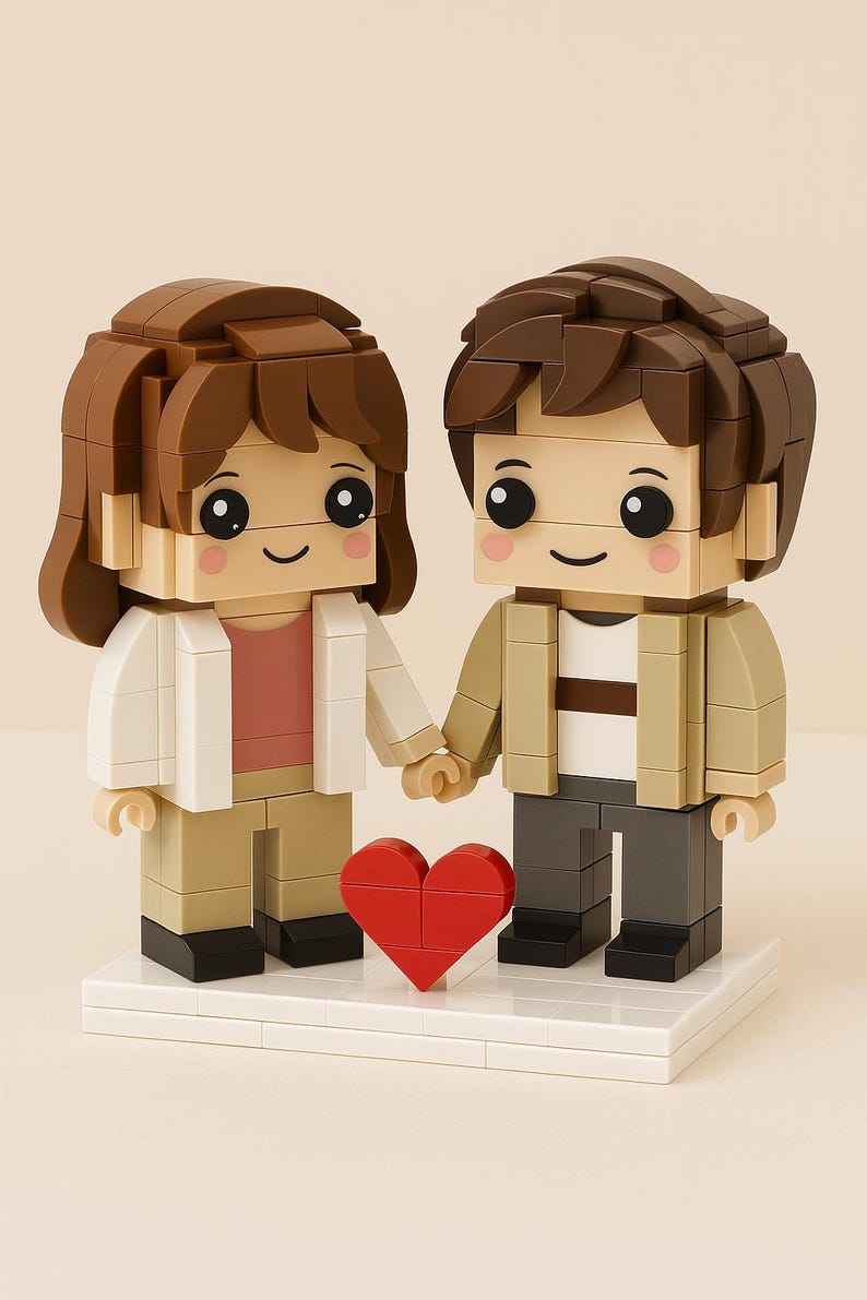 May include: A Lego-style couple figurine holding hands, with a red heart between them. The figures have brown hair, smiling faces, and are dressed in neutral-toned clothing. The base is white.