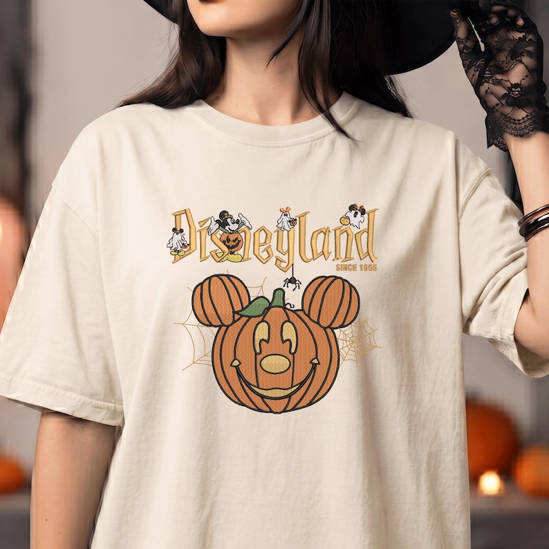 May include: A cream-colored t-shirt featuring a graphic of a pumpkin shaped like Mickey Mouse ears. Above the pumpkin, the text reads Disneyland SINCE 1955, accompanied by small illustrations of ghosts and a spider. The design includes spiderwebs.