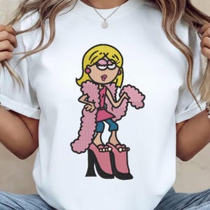 May include: White t-shirt featuring an embroidered cartoon character in a pink outfit, blue jeans, and pink high heels. The character has blonde hair, glasses, and a pink boa. The design is a playful, cartoon-style illustration.