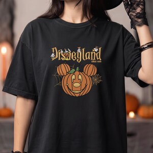 May include: A black short-sleeve t-shirt featuring a graphic of a pumpkin shaped like a mouse head with ears, surrounded by spider webs. The text Disneyland Since 1955 is printed above the pumpkin in a gold font, with small ghost illustrations.