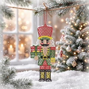 May include: A hanging nutcracker ornament featuring a red, green, and yellow honeycomb-patterned design. The figure wears a tall hat, holds a red and green gift box, and has a black mustache. It is suspended by a thin red string from a branch.