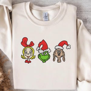May include: Cream-colored sweatshirt featuring embroidered cartoon faces of Cindy Lou Who, the Grinch, and Max, each wearing a Santa hat. The Grinch is green, Cindy Lou Who is blonde, and Max is brown. A festive holiday-themed design.