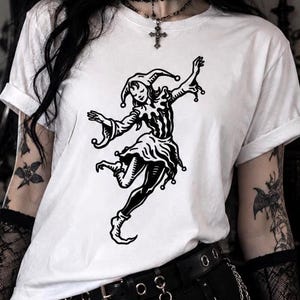 May include: A white short-sleeved t-shirt featuring a black graphic print of a jester in a dynamic dancing pose. The shirt has a crew neckline and cuffed sleeves, showcasing a detailed illustration of a jester wearing a traditional cap with bells.