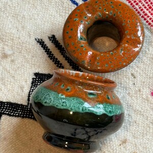 Puede incluir: Dos piezas de cerámica: un jarrón pequeño y redondo y un objeto en forma de anillo. El jarrón es verde oscuro en la base, con una banda turquesa y una parte superior naranja. El anillo es naranja con motas turquesas.