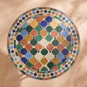 Op de afbeelding: Een rond tafelblad met een geometrisch mozaïekpatroon bestaande uit achthoekige en ruitvormige tegels in tinten blauw, groen, geel, oranje en wit. Het oppervlak is gerangschikt in een herhalend symmetrisch ontwerp met een donkere rand.