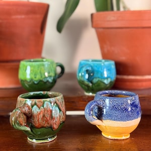 May include: Four small, handmade ceramic mugs with unique, colorful glazes. The mugs feature a variety of colors, including green, blue, and a mix of brown and red. Each mug has a small handle and sits on a wooden surface.