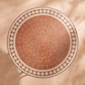 Puede incluir: Un tablero de mesa redondo con una superficie de mosaico de color terracota dispuesta en un patrón geométrico de estrella en el centro, rodeado por círculos concéntricos de azulejos y un borde exterior de rombos blancos y terracota.