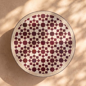 Op de afbeelding: Een rond tafelblad met een geometrisch mozaïekpatroon van diepbordeauxrode en crèmekleurige tegels. Het ontwerp bestaat uit herhalende achtpuntige sterren en kleine vierkantjes, omrand door een rand van rechthoekige crèmekleurige tegels.