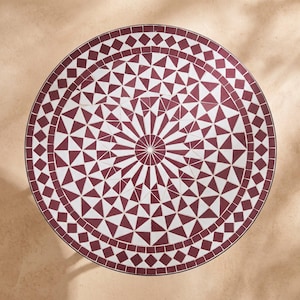 Op de afbeelding: Een ronde tafel met een mozaïekblad met een geometrisch patroon van donkerrode en witte tegels. Het ontwerp bestaat uit een centraal stervormig motief omringd door concentrische ringen van driehoeken en ruiten, afgewerkt met een donkere rand.