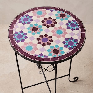 Può includere: Un tavolino rotondo con piano in mosaico caratterizzato da un motivo geometrico a stella nei toni del viola, rosa e turchese. Il tavolo presenta una sottile struttura in metallo nero con quattro gambe slanciate e un supporto a crociera.