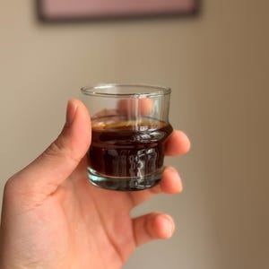 Può includere: Un piccolo bicchierino di vetro trasparente riempito con un liquido marrone scuro, probabilmente caffè o espresso. Il bicchiere è tenuto in una mano, con il liquido visibile. Il bicchierino ha una base spessa e un bordo leggermente svasato. Lo sfondo è sfocato.