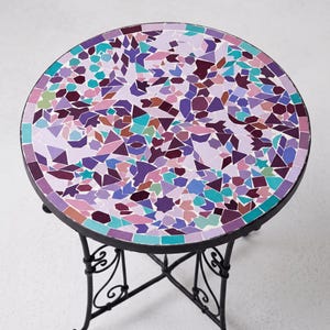 Peut inclure: Une table ronde avec un plateau en mosaïque composé de carreaux irréguliers dans des tons de violet, lavande, magenta, bleu sarcelle et vert clair. La table repose sur une base en métal noir avec quatre pieds incurvés et une entretoise.