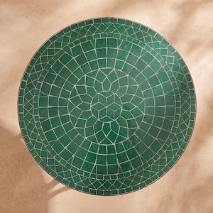 Peut inclure: Un plateau de table circulaire présentant un motif de mosaïque verte disposé en formes géométriques concentriques. La surface se compose de nombreuses petites tuiles vertes de formes irrégulières séparées par de fines lignes de joint.