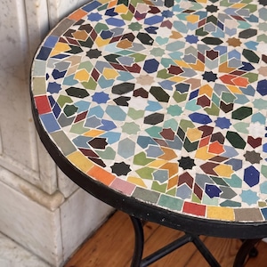 Peut inclure: Une table ronde avec un plateau en mosaïque au motif géométrique en étoile, composé de petites tuiles multicolores dans des tons de bleu, jaune, vert, rouge et blanc, reposant sur un cadre fin en métal noir aux pieds incurvés.