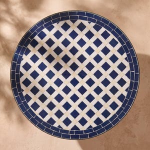 Peut inclure: Un plateau de table circulaire présentant un motif en mosaïque composé de carreaux carrés bleu foncé et blanc cassé disposés en grille en losange. Le bord extérieur est bordé d'une rangée de carreaux rectangulaires bleu foncé.