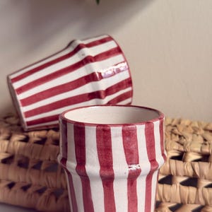 Peut inclure: Deux tasses en céramique à rayures rouges et blanches. L'une est debout, l'autre est couchée. Les tasses ont une forme évasée et une surface texturée. L'arrière-plan comprend un panier tressé et de la verdure.