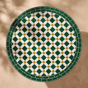 May include: A round table top featuring a mosaic pattern of dark green, white, and mustard yellow tiles. The design consists of a central grid of diamonds with alternating colors, surrounded by a solid dark green border of rectangular tiles.