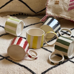 Peut inclure: Plusieurs tasses en céramique avec des motifs à rayures colorées. Les tasses présentent des rayures vertes, rouges, jaunes, noires et vertes. Chaque tasse a une poignée assortie. Les tasses sont présentées sur un tapis à motifs.