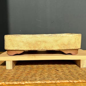 Puede incluir: Una maceta rectangular de cerámica amarillo claro con superficie texturizada. La maceta tiene una base marrón rojizo y se apoya en un soporte de bambú. Diseñada para cultivar árboles y plantas pequeñas.