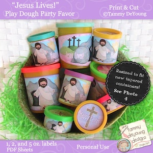 May include: A set of colourful play dough containers with labels featuring a religious Easter theme. The labels include images of Jesus, crosses, and the text "Jesus Lives!" and "Happy Easter!" The containers are in a basket filled with green grass.