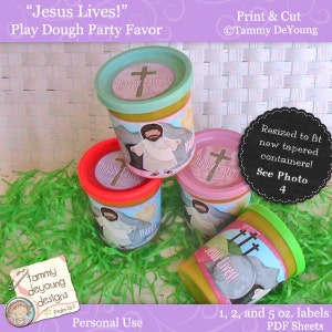 May include: Four colourful play dough containers with Easter-themed labels. The labels feature a cross, the words "Happy Easter", and "Jesus Lives!" The containers are sitting on a bed of green grass.
