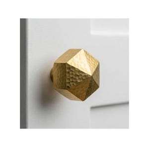 May include: A faceted, gold-colored door knob with a hammered texture. The knob is mounted on a white door, showcasing its geometric design and metallic finish. This decorative hardware adds a touch of elegance to any interior.