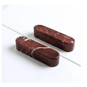 Könnte beinhalten: Zwei rechteckige Schrankgriffe aus rotbraunem Marmor mit weißen Adern. Die Griffe haben abgerundete Kanten und werden auf einer weißen Oberfläche präsentiert. Diese dekorativen Griffe sind ideal, um Möbeln einen Hauch von Eleganz zu verleihen.