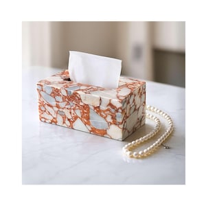 May include: A rectangular tissue box made of marble with red, white, and gray patterns. A white tissue is pulled from the top opening. A pearl necklace rests on the surface next to the box.