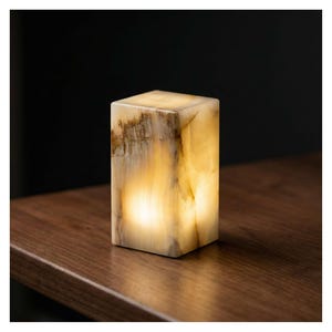 May include: A rectangular, illuminated onyx lamp. The lamp has a warm, yellow glow emanating from within, with brown and beige veining on the exterior. It sits on a dark wooden surface, with a black background.