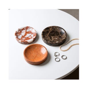May include: Three small, round marble dishes in shades of red and white, brown and black, and orange. A pearl necklace and three rings are next to the dishes. The dishes are on a white surface.