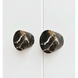 Peut inclure: Deux boutons de meuble cylindriques en marbre noir avec veinage doré et blanc. Les boutons sont montés sur une porte d'armoire blanche, offrant une esthétique moderne et élégante. Ces boutons décoratifs sont parfaits pour moderniser les meubles.