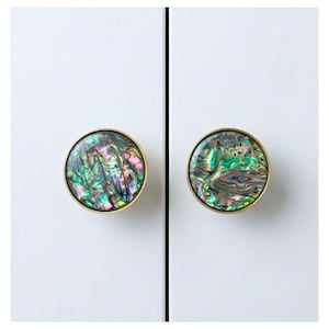 May include: Two round cabinet knobs with iridescent abalone shell centers and gold-colored metal rims. The knobs are mounted on a white cabinet door. The abalone shell displays a range of colors, including green, blue, and purple.
