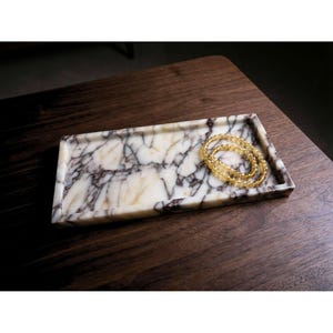 Könnte beinhalten: Ein rechteckiges Marmortablett mit einem cremefarbenen und braunen Muster. Ein goldfarbenes Perlenarmband liegt in der Schale. Das Tablett steht auf einer dunkelbraunen Holzoberfläche. Dieses Tablett eignet sich für Schmuck oder kleine Gegenstände.