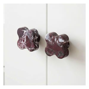 Könnte beinhalten: Zwei dekorative Schrankknöpfe mit einem vierblättrigen Kleeblatt-Design. Die Knöpfe sind aus dunkelrotem und weißem Marmor gefertigt. Sie sind an einer weißen Schranktür montiert.