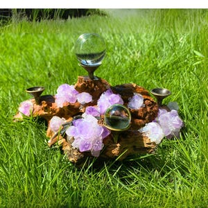 May include: A decorative crystal display featuring a piece of driftwood adorned with clusters of purple amethyst crystals and clear glass spheres. The display also includes small, dark metal candle holders. The arrangement is set against a backdrop of green grass.