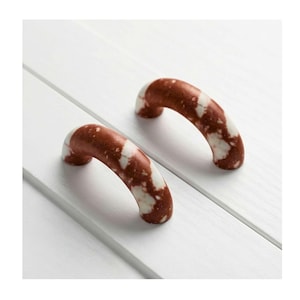 May include: Two curved cabinet pulls with a brown and white speckled pattern. The pulls are arched and appear to be made of a solid material. They are set against a white wooden surface.