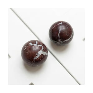 May include: Two round, marble spheres with a deep red-brown hue and white veining. The spheres are positioned on a white surface, showcasing their polished texture and natural patterns. The contrasting colors create a striking visual.