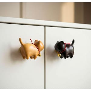 May include: Two cat-shaped cabinet knobs. One is light brown, the other is black. Each has a red collar with a small gold bell. The knobs are mounted on a white cabinet door. These decorative knobs add a whimsical touch.