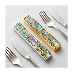 May include: Two rectangular abalone shell knife rests, one gold-toned and one silver-toned, are displayed with stainless steel forks and knives. The iridescent shell pattern features shades of green, blue, and purple. The cutlery is arranged on a white surface.