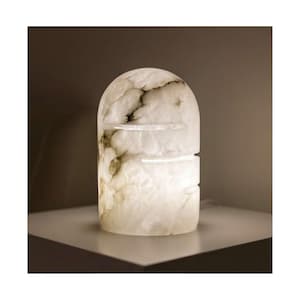 Könnte beinhalten: Eine weiße, bogenförmige Tischlampe mit marmorähnlichem Aussehen. Die Lampe hat einen rechteckigen Sockel und eine abgerundete Oberseite mit dunklen Adern. Sie hat zwei horizontale Aussparungen und strahlt ein weiches, warmes Licht aus.
