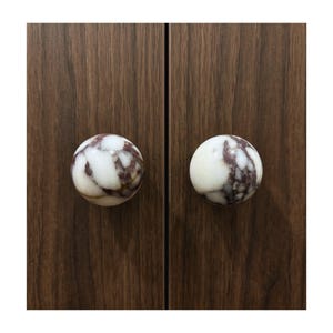 May include: Two round cabinet knobs with a white and brown marbled pattern. The knobs are attached to a dark brown wooden cabinet door. The knobs are a decorative element for furniture.