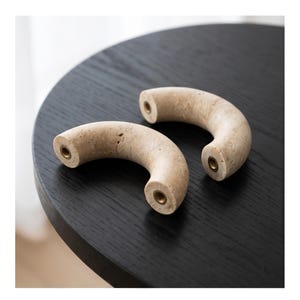 May include: Two curved, beige stone cabinet pulls with brass inserts. The pulls are in a half-moon shape and rest on a dark, round wooden table. The stone has a natural, porous texture. These decorative pulls are ideal for adding a unique touch to furniture.
