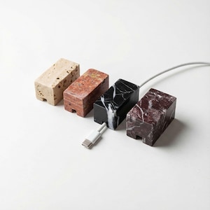 May include: Four rectangular cable tidies in beige, red, black with white veining, and maroon stone. A white USB-C cable is connected to the black tidy. The tidies are designed to hold and manage cables, keeping them neat.