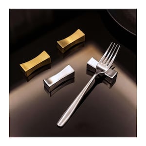 May include: A stainless steel fork rests on a silver fork rest. Two gold and one silver fork rests are arranged on a reflective black surface. The fork rests have a curved, rectangular design, and the fork is sleek and modern.