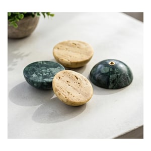 May include: Four decorative stone bottle stoppers in shades of green and tan. Two are a mottled green, one is tan with small holes, and one is a darker green with a gold accent. The stoppers are round and sit on a light-colored surface.