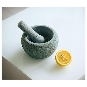 Puede incluir: Un juego de mortero y maja de piedra gris con una superficie texturizada. Un limón amarillo cortado por la mitad está al lado del mortero. El mortero y la maja están sobre una superficie blanca.